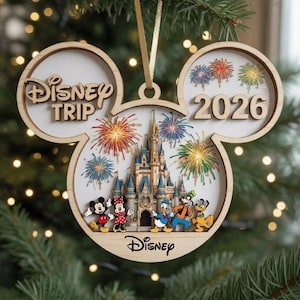 May include: A wooden Mickey Mouse-shaped Christmas ornament featuring a castle, fireworks, and Disney characters. The text reads Disney Trip and 2026. The ornament has a light wood frame and a gold ribbon for hanging, showcasing a colourful scene.