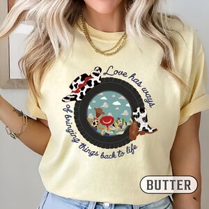 May include: A pale yellow t-shirt featuring a graphic of a black tire framing a sky-blue background with cartoon characters. The text reads, Love has ways of bringing things back to life, accompanied by a cow-print hat and a cow-print cowboy boot.