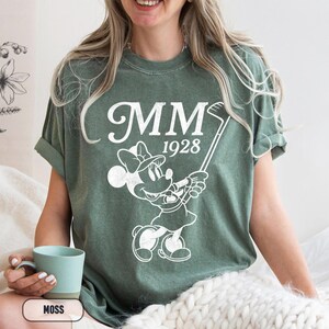 Puede incluir: Camiseta verde musgo con un gr&aacute;fico blanco de Minnie Mouse jugando al golf, con el texto "MM 1928". La camiseta es de corte holgado y tiene mangas enrolladas. Una persona sostiene una taza turquesa.