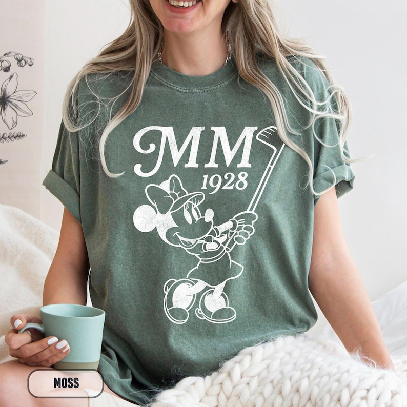 Puede incluir: Camiseta verde musgo con un gr&aacute;fico blanco de Minnie Mouse jugando al golf, con el texto "MM 1928". La camiseta es de corte holgado y tiene mangas enrolladas. Una persona sostiene una taza turquesa.