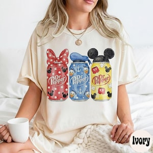Pode incluir: Camiseta bege com três latas de Dr. Pepper com designs da Disney. As latas são vermelhas com um laço da Minnie Mouse, azuis com o Pato Donald e amarelas com orelhas do Mickey Mouse. O logotipo da Dr. Pepper é visível em cada lata.