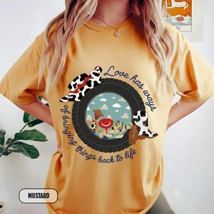 May include: A mustard yellow short-sleeve t-shirt featuring a graphic of a black tire surrounding a sky-blue background with cartoon characters. The text "Love has ways of bringing things back to life" curves around the tire, with cow-print boots.