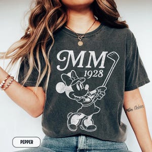 Puede incluir: Camiseta gris oscuro con un gr&aacute;fico blanco de Minnie Mouse jugando al golf, con el texto "MM 1928". La camiseta tiene un aspecto vintage. La palabra "PEPPER" est&aacute; en un cuadro blanco en la parte inferior.