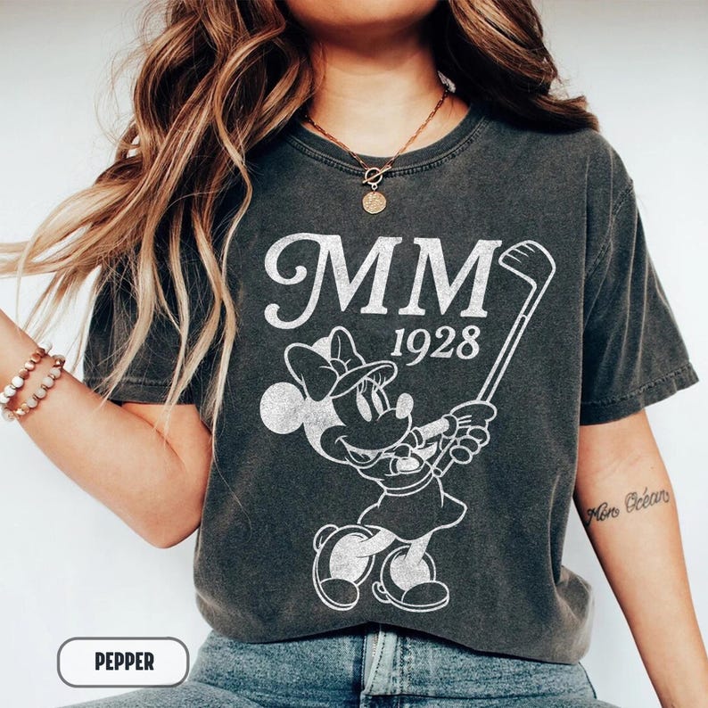 Puede incluir: Camiseta gris oscuro con un gr&aacute;fico blanco de Minnie Mouse jugando al golf, con el texto "MM 1928". La camiseta tiene un aspecto vintage. La palabra "PEPPER" est&aacute; en un cuadro blanco en la parte inferior.