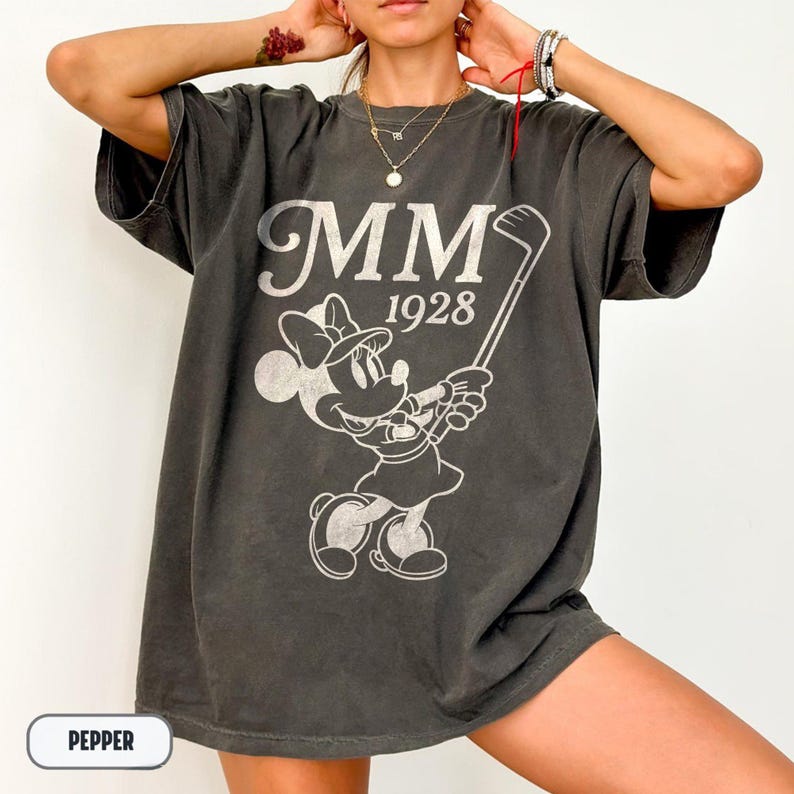 Puede incluir: Camiseta gris oscuro con un gr&aacute;fico blanco de Minnie Mouse jugando al golf, con el texto "MM 1928". La camiseta tiene un corte holgado y mangas cortas. La palabra "PEPPER" est&aacute; en un cuadro blanco en la parte inferior.