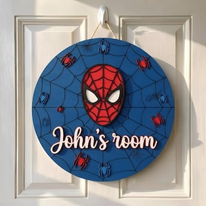 Könnte beinhalten: Ein rundes Türschild aus Holz, blau lackiert mit schwarzem Spinnennetz-Muster. In der Mitte befindet sich ein roter und schwarzer Maskenausschnitt. Der Text John's room ist in weißer Schrift zu sehen. Kleine rote und blaue Spinnen sind befestigt.