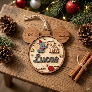 May include: A wooden ornament shaped like a mouse head featuring a layered design with a Dumbo elephant character, a circus tent, clouds, stars, and a red ball. The text reads 2026, 1st Christmas, and the name Lucas in a dark script font.