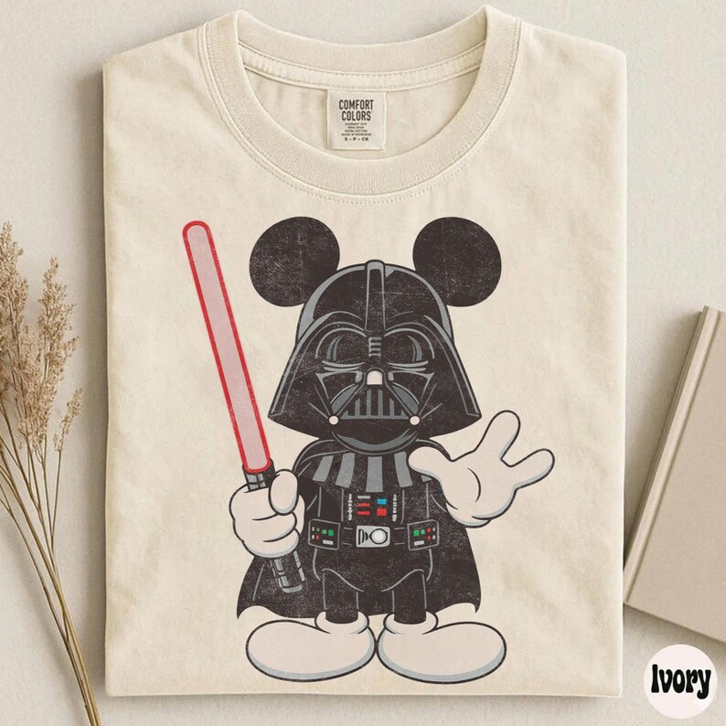 Pu&ograve; includere: T-shirt color avorio con un personaggio dei cartoni animati di Darth Vader con le orecchie di Topolino, che tiene una spada laser rossa. Il personaggio indossa un casco e un mantello neri. La maglietta ha un'etichetta Comfort Colors.