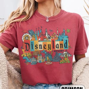 K&ouml;nnte beinhalten: Karmesinrotes T-Shirt mit einem farbenfrohen Disneyland Resort-Grafik. Das Design zeigt ikonische Parkbilder und das Disneyland-Logo in einem Retro-Stil. Das Shirt ist bequem und l&auml;ssig.