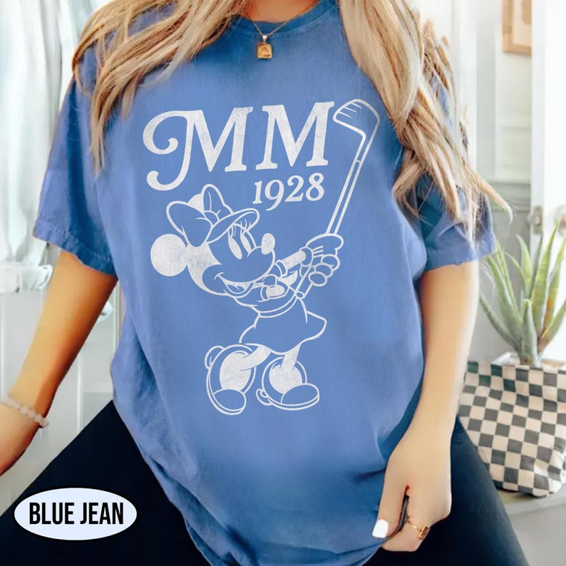 Puede incluir: Camiseta azul con un gr&aacute;fico blanco de Minnie Mouse jugando al golf, el texto "MM 1928" est&aacute; encima del personaje. La camiseta tiene mangas cortas y un corte holgado. El texto "BLUE JEAN" est&aacute; en la parte inferior.