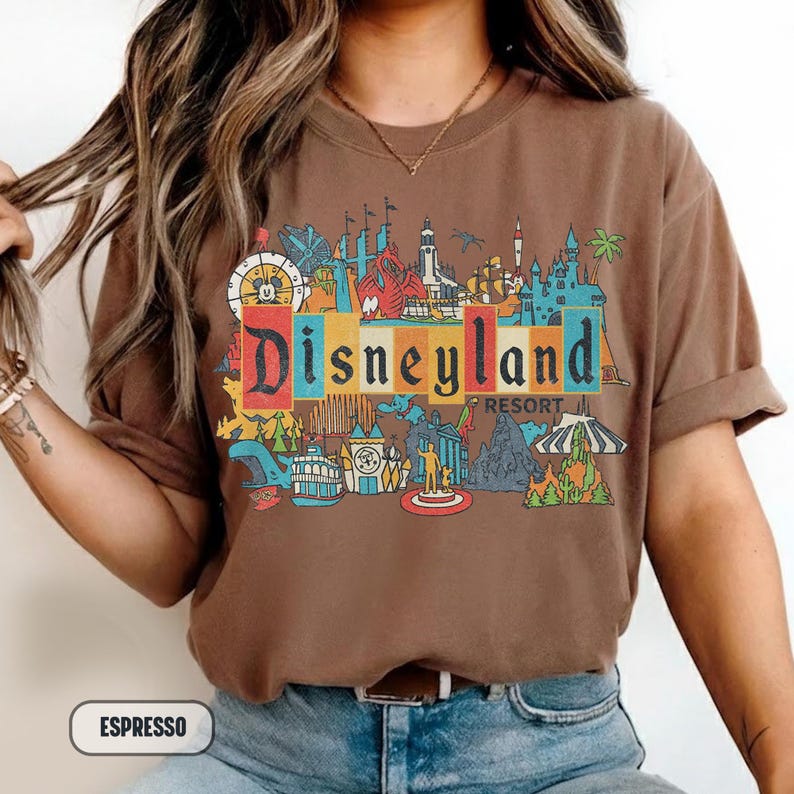 K&ouml;nnte beinhalten: Espressofarbenes T-Shirt mit einem farbenfrohen Disneyland Resort-Motiv. Das Design beinhaltet das Disneyland-Logo und verschiedene Parksymbole. Das Shirt hat kurze &Auml;rmel und einen Rundhalsausschnitt.