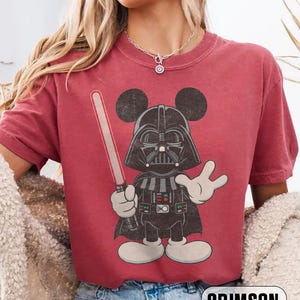 Pu&ograve; includere: T-shirt color cremisi con un cartone animato che combina Darth Vader e Topolino. Il personaggio tiene una spada laser rossa e indossa un casco nero con le orecchie di Topolino. La parola "CRIMSON" &egrave; stampata in basso.