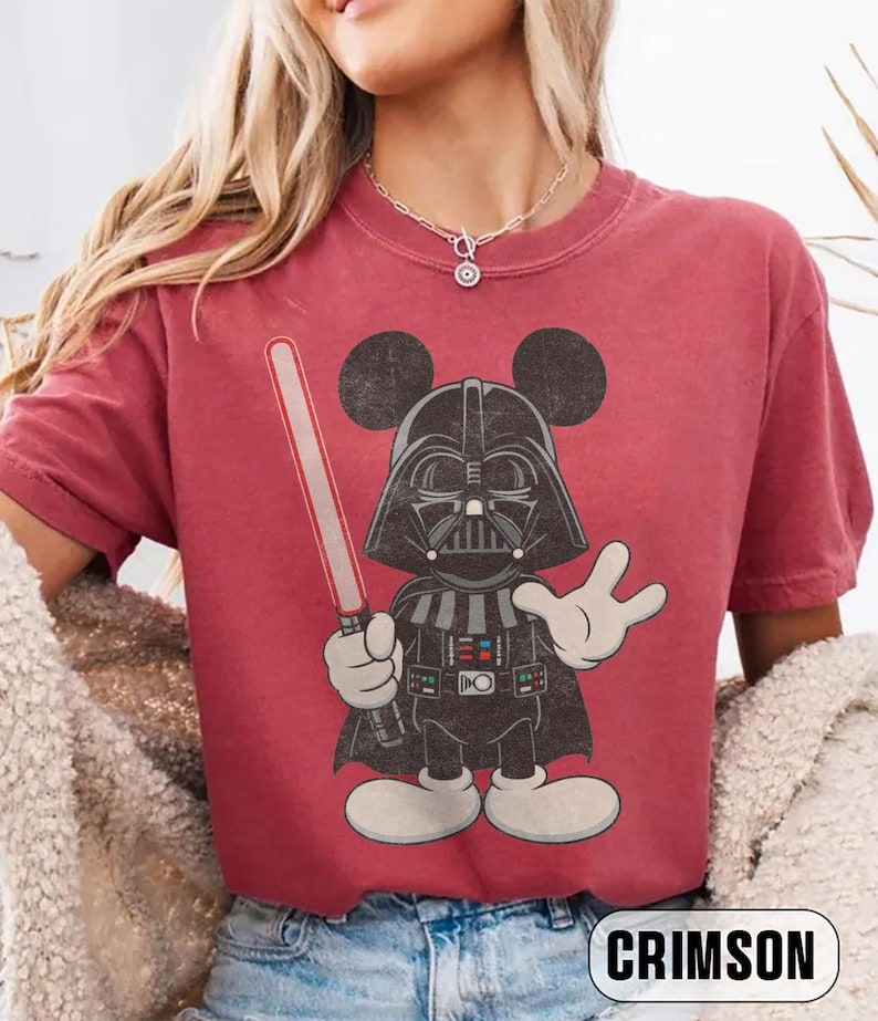 Pu&ograve; includere: T-shirt color cremisi con un cartone animato che combina Darth Vader e Topolino. Il personaggio tiene una spada laser rossa e indossa un casco nero con le orecchie di Topolino. La parola "CRIMSON" &egrave; stampata in basso.