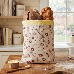 May include: A beige bread bag with a floral pattern and the text "Fresh Bread Bag" holds two loaves of bread. One is round with a dark crust, the other is a braided loaf with sesame seeds. A small dish of butter and a wooden knife are on the table.