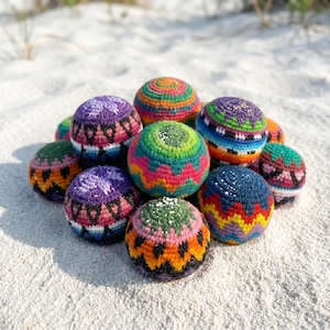 May include: A collection of colorful, hand-crocheted balls in various sizes. The balls feature vibrant patterns in shades of purple, green, orange, blue, and pink. They are arranged on a sandy surface, suggesting a beach or outdoor setting.