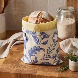 Puede incluir: Una hogaza de pan redonda dentro de una bolsa de pan floral. La bolsa es de color crema con patrones florales azules y un interior amarillo. La bolsa está sobre una tabla de cortar de madera.