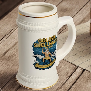 Może przedstawiać: Biały ceramiczny kufel do piwa z uchwytem, ze złotą obwódką i dekoracyjnym wzorem. Wzór zawiera napis "GOLDEN SHELLBACK US NAVY" oraz ilustrację mężczyzny w oceanie. Kufel stoi na drewnianym stole.