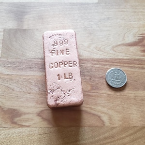 May include: A rectangular copper bar stamped with "999 Fine Copper 1 LB" next to a quarter coin. The copper bar has a textured surface and a reddish-brown color, set on a wooden surface.