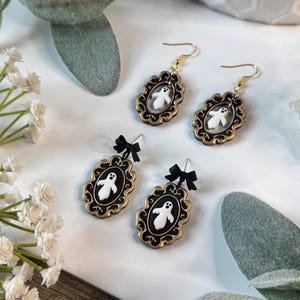Op de afbeelding: Vier paar Halloween-oorbellen. Twee paar hebben goudkleurige frames met een zwart interieur en een wit spookontwerp. De andere twee paar hebben zwarte strikken. De oorbellen worden gepresenteerd op een wit oppervlak met decoratief groen.