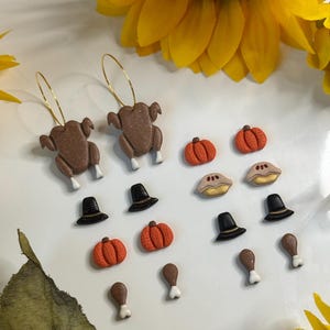 May include: Thanksgiving-themed earrings and charms. The earrings feature brown turkey pendants on gold hoops. Charms include orange pumpkins, pilgrim hats, pie slices, and turkey legs. The items are arranged on a white surface with a sunflower in the background.