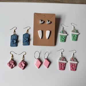 May include: A collection of handmade earrings in various designs. Includes blue rectangular earrings with floral accents, heart-shaped studs with polka dots, and pink flower earrings. Also, pink diamond-shaped and bubble tea earrings.
