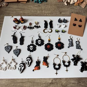 Könnte beinhalten: Eine Sammlung handgefertigter Halloween-Ohrringe. Die Ohrringe zeigen verschiedene Designs, darunter Geister, Fledermäuse, Totenköpfe und Kürbisse. Die Farbpalette umfasst Schwarz, Weiß, Orange und Gold. Einige Ohrringe sind Stecker, andere sind Hängeohrringe.