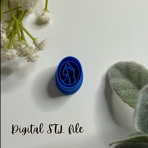 May include: A blue plastic oval-shaped cookie cutter with a silhouette design of a woman's profile. The text "Digital STL file" is written in black cursive. White flowers and green leaves are in the background.