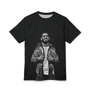 May include: Black t-shirt featuring a grayscale image of a man holding two championship belts. The image is a portrait of a smiling man wearing a white shirt and a gray hoodie.