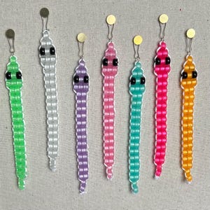 May include: Seven colorful beaded snake keychains with black eyes. The snakes are made of small, round beads in various colors, including green, white, purple, pink, turquoise, and orange.