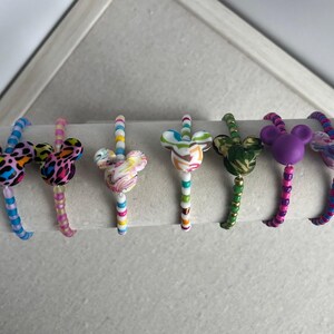 May include: A collection of eight colorful bracelets, each featuring a Mickey Mouse head charm. The charms display various patterns and colors, including leopard print, marble, and solid hues. The bracelets are displayed on a white cylindrical stand.
