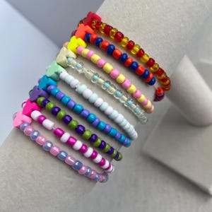 May include: A collection of beaded bracelets in various colors, including red, orange, yellow, blue, green, purple, pink, and white. Each bracelet features a heart-shaped bead at the end. The bracelets are displayed on a white cylindrical surface.
