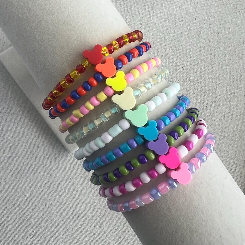 May include: A collection of beaded bracelets in various colors, each featuring a Mickey Mouse-shaped charm. The bracelets are arranged in a layered display, showcasing a range of hues from red and yellow to blue, green, and pink. The beads are small and round.