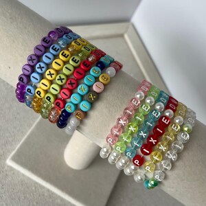 May include: A collection of colorful beaded bracelets. Each bracelet features the word "PIXIE" spelled out with letter beads. The bracelets are in various colors, including purple, blue, yellow, red, and pink. Some bracelets also have the word "EDU".