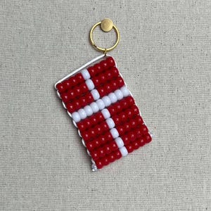 May include: A red and white beaded keychain shaped like the flag of Denmark. The keychain has a gold metal ring.