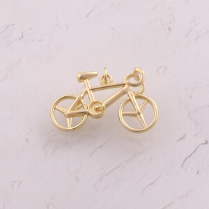 May include: A gold-colored bicycle charm with a detailed design. The charm features a classic bicycle frame, wheels, seat, and handlebars. The bicycle charm is made of a shiny, metallic material and is likely a jewelry component or decorative item.