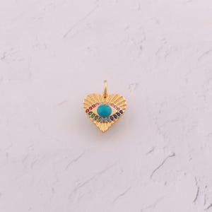 May include: A gold-colored heart-shaped pendant with a turquoise eye design. The eye is surrounded by a rainbow of colorful gemstones. The pendant has a textured, sunburst-like pattern around the eye.