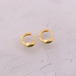 May include: Two gold-colored hoop earrings with a small, green gemstone set into each. The earrings are round with a hinged closure. The background is a light, textured surface.
