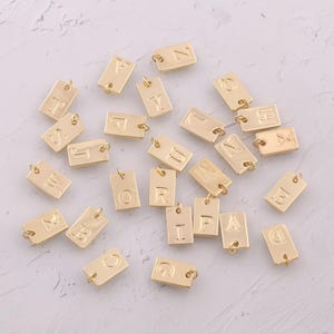 May include: Gold-colored rectangular charms with various engraved letters. Each charm has a small loop at the top for attaching to jewelry. The charms are scattered on a light-colored surface, showcasing a variety of alphabet letters.