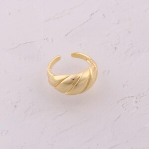 Puede incluir: Un anillo dorado con un diseño texturizado y acanalado. El anillo tiene un diseño abierto y ajustable. El anillo está sobre un fondo de mármol blanco y gris. El anillo es una joya.