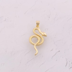 Peut inclure: Pendentif doré en forme de serpent. Le serpent est enroulé, avec une tête et un corps détaillés. Il est suspendu à une petite boucle. Le fond est une surface claire et mouchetée.