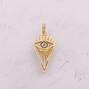Puede incluir: Colgante dorado en forma de ojo, con base triangular. El iris del ojo es negro, rodeado de cristales blancos y negros. El colgante está adornado con cristales de colores y un diseño de rayos de sol.