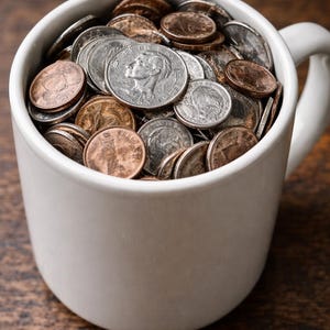 Könnte beinhalten: Eine weiße Keramiktasse, gefüllt mit einer Vielzahl von Münzen. Die Münzen sind in Silber- und Kupfertönen gehalten, einige zeigen Porträts. Die Tasse steht auf einer Holzoberfläche.