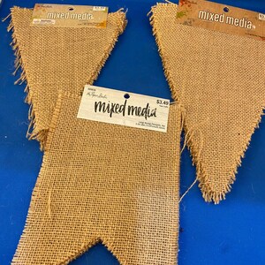 May include: Three burlap pennants with frayed edges. Each pennant has a tag that reads "mixed media". The pennants are a natural tan color and are arranged on a blue surface.