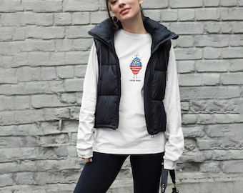 Op de afbeelding: Een wit shirt met lange mouwen met een cartoon uil ontwerp in rood, wit en blauw. De tekst "FREE BIRD" staat onder de uil gedrukt. Het shirt wordt gedragen met een zwart vest en leggings.