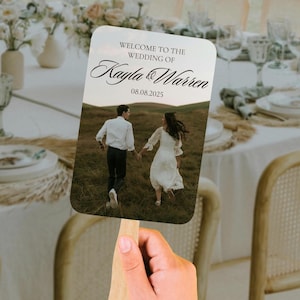 May include: A wedding fan with a photo of a couple running through a field, held by a hand. The fan has text that reads "Welcome to the Wedding of Kayla & Warren 08.08.2025". The fan is made of paper and wood.