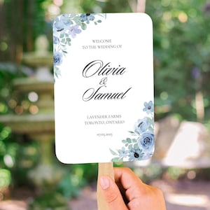 May include: A white wedding fan with a wooden handle, held by a hand. The fan features floral accents in shades of blue and the text "Welcome to the Wedding of Olivia & Samuel." The wedding date is 07.05.2025.