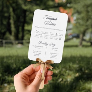 Peut inclure: Un éventail de mariage blanc avec un ruban doré, portant les noms "Hannah & Walter" et un programme de mariage. L'éventail énumère les membres du cortège nuptial, y compris la demoiselle d'honneur, les demoiselles d'honneur, le témoin et les garçons d'honneur.