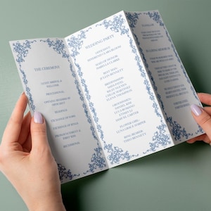 May include: A folded wedding program with blue floral border designs and text. The program lists the wedding party, ceremony details, and a memorial section. The program is held open by two hands.