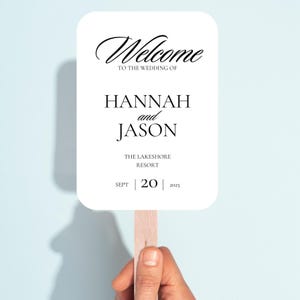 May include: A white wedding fan with rounded corners, featuring the words "Welcome" and "To the wedding of HANNAH and JASON". The date "SEPT 20 2025" is printed at the bottom. The fan is held by a wooden stick.