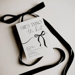 May include: A white rectangular bridal shower invitation card featuring black text that reads "SHE'S TYING the knot" above a black line drawing of a bow. The card includes event details and is accented by a long, flowing black satin ribbon.